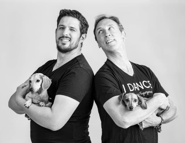 Half body shot of two smiling men back to back holding mini dachshunds
